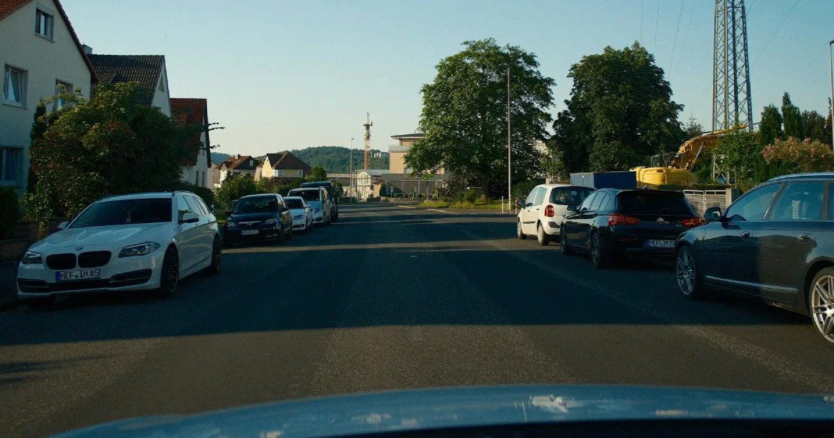 Residential street with parked cars and a transmission tower rising behind the houses.