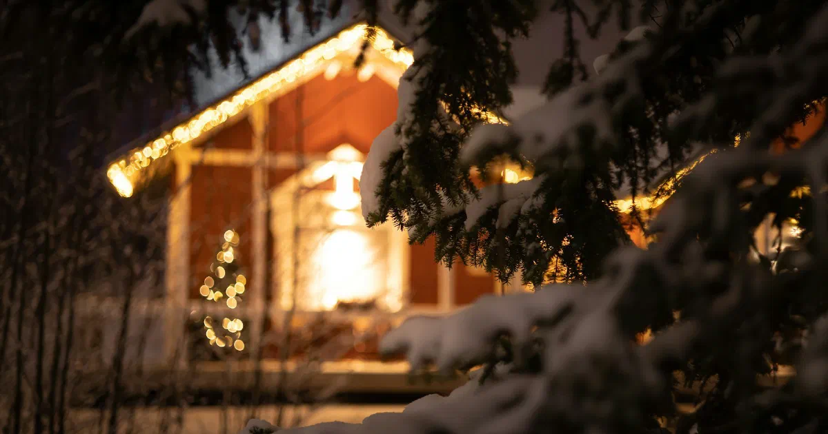 House glowing with warm lights at night behind snow-covered evergreen branches.