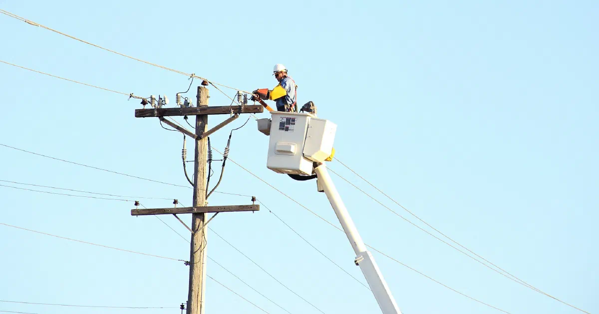 Utility worker in a bucket lift working on a power pole against a clear sky.