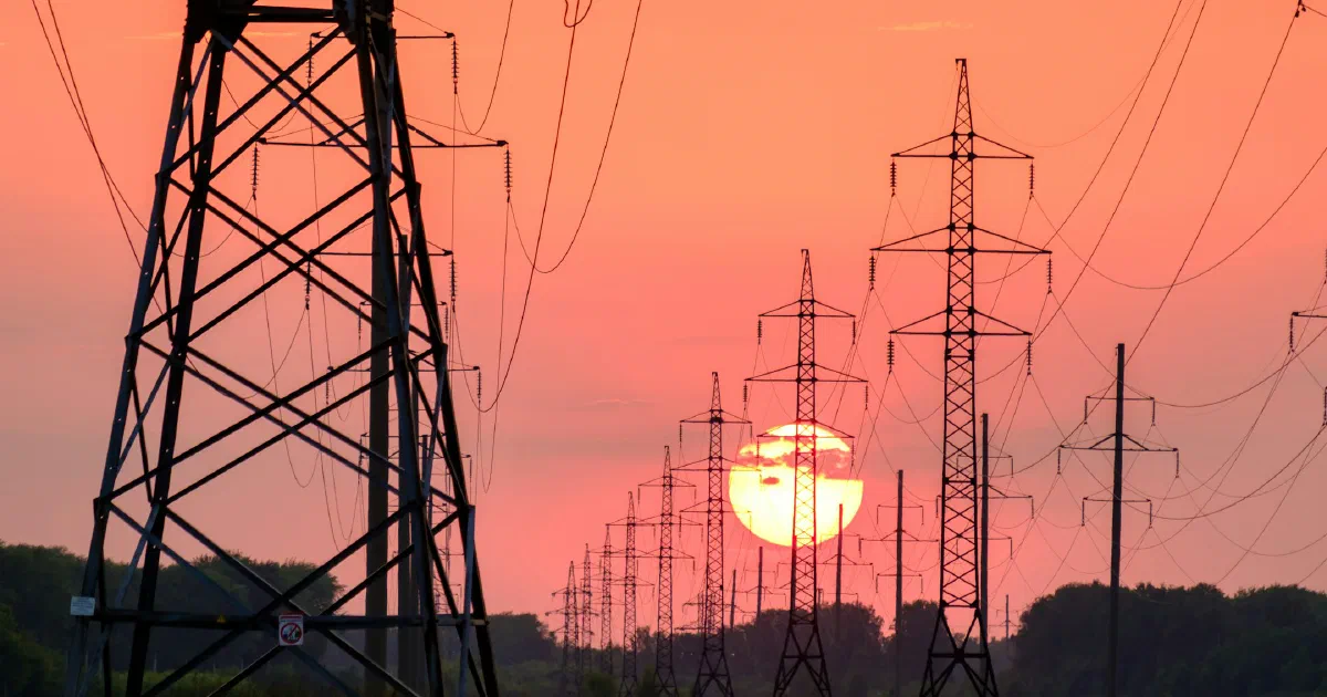 Silhouetted transmission towers and power lines stretching toward a setting sun.