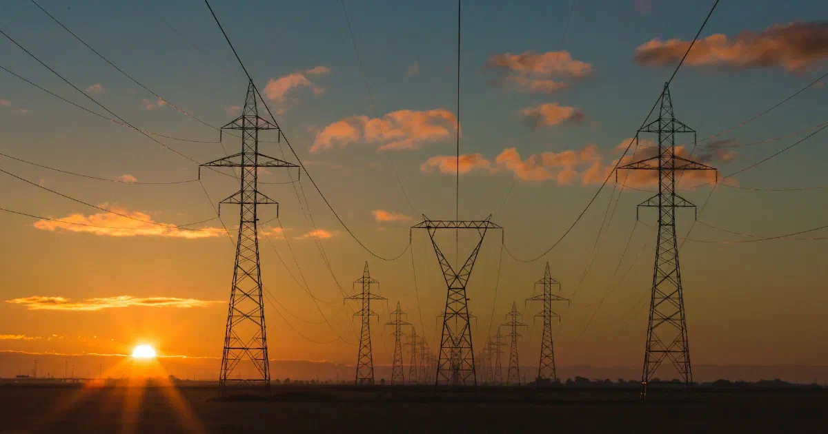 Rows of transmission towers and power lines silhouetted against an orange sunset.