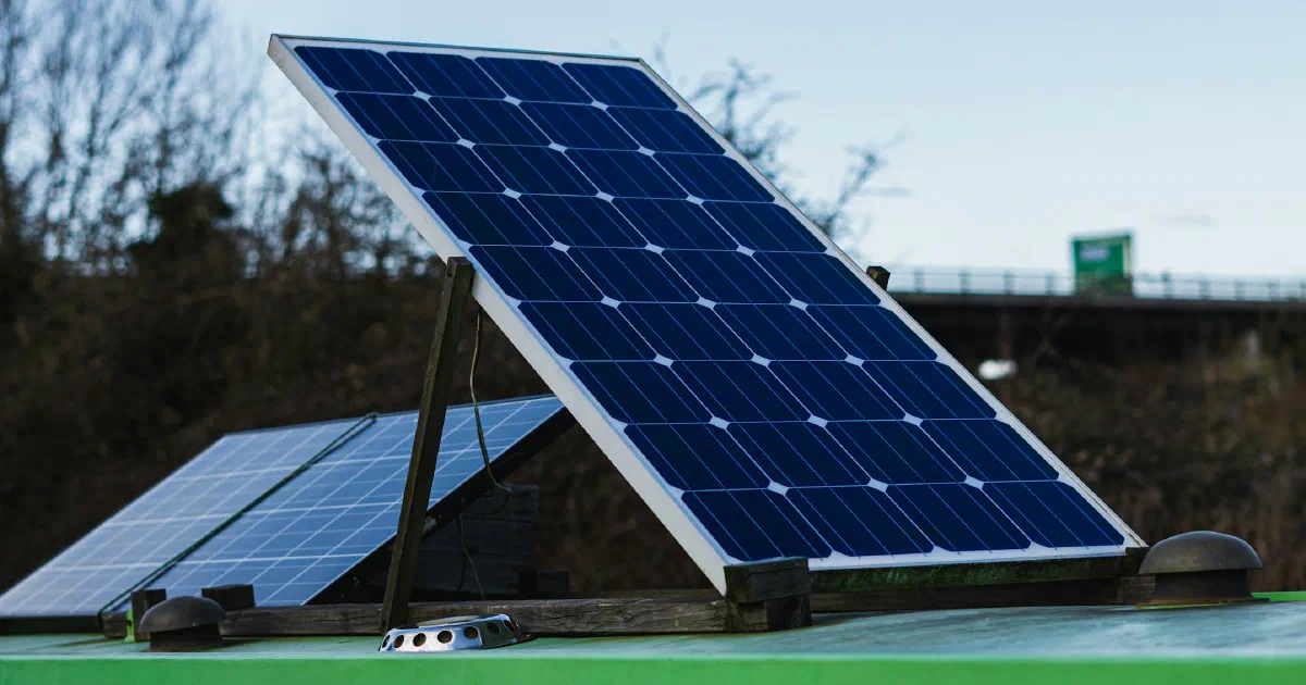 A solar panel tilted on a mount above a green surface with bare trees behind it.