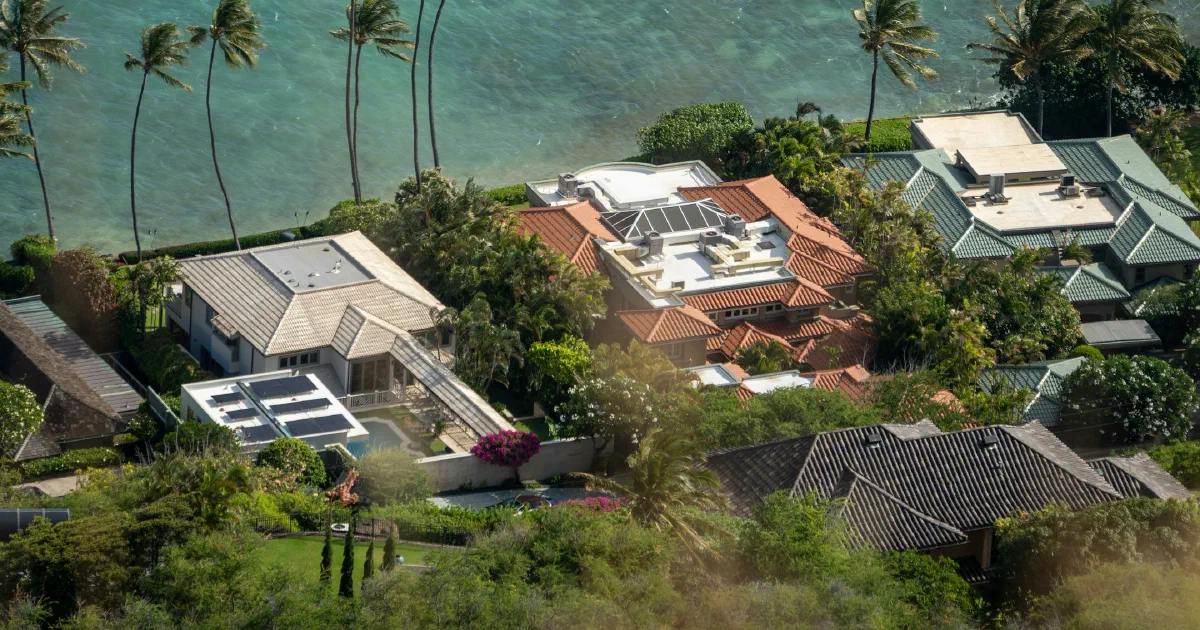Aerial view of waterfront houses with palm trees beside turquoise water, some roofs fitted with solar panels.