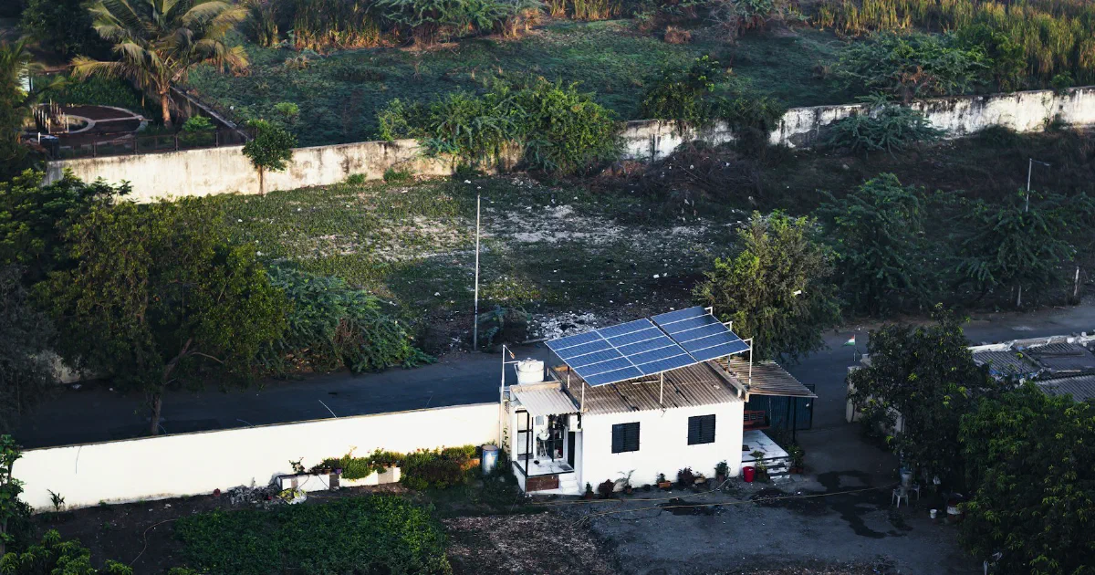 Small white house with rooftop solar panels surrounded by tropical trees and garden plots.