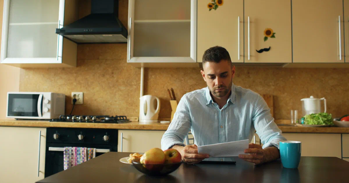 Man sitting at a kitchen table reading a paper bill, with a cup and a bowl of fruit in front of him.