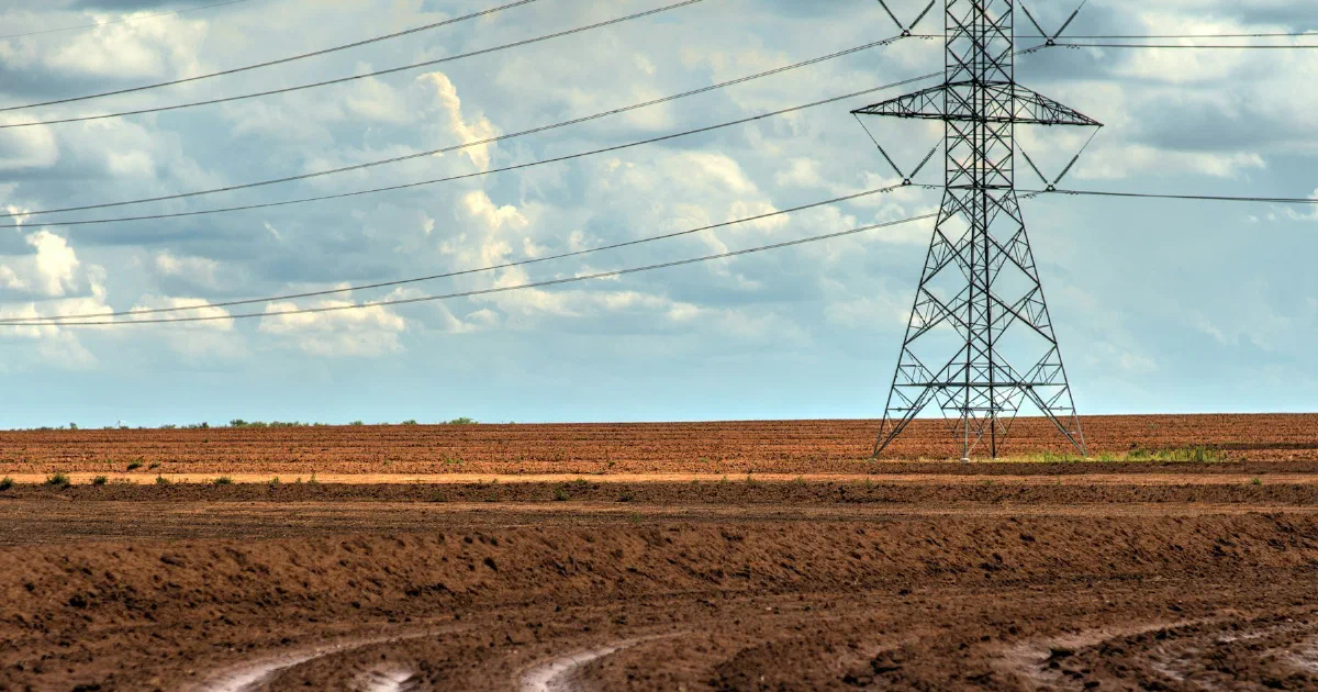 Steel transmission tower and power lines above a plowed field under a cloudy sky.