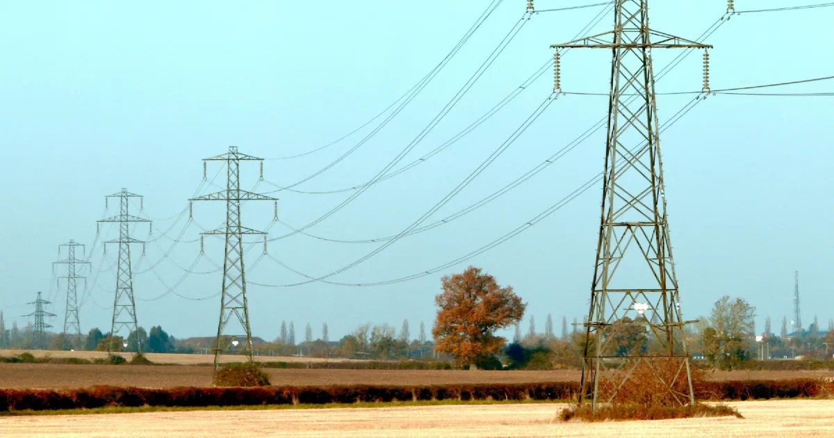 Line of transmission towers crossing a harvested field under a pale blue sky.