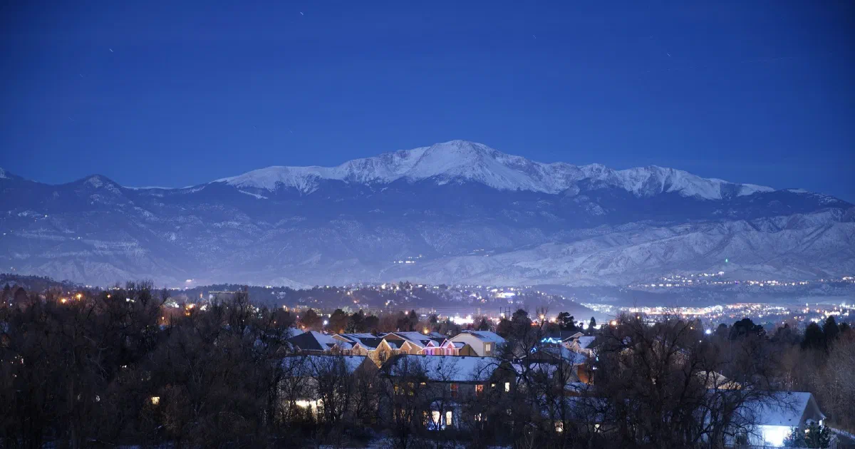 Snowy neighborhood at dusk with lit windows below a snow-capped mountain.