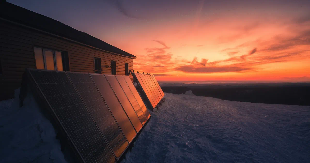 Solar panels on a roof edge in the snow, lit by an orange sunset.