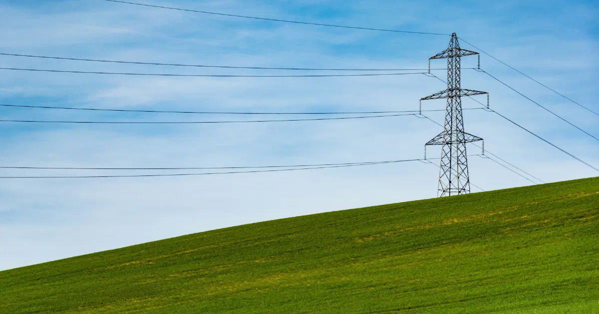 Transmission tower and power lines above a green hillside under a blue sky with thin clouds.