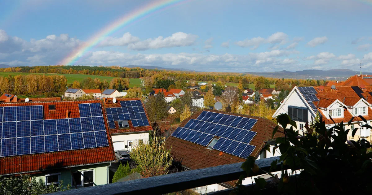 Rooftop solar panels in a residential neighborhood with a rainbow over hills behind.