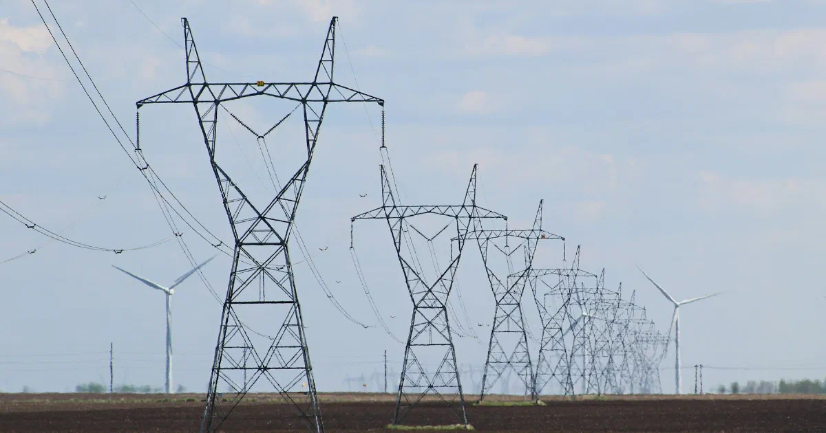 Transmission towers crossing a flat field with wind turbines in the distance.