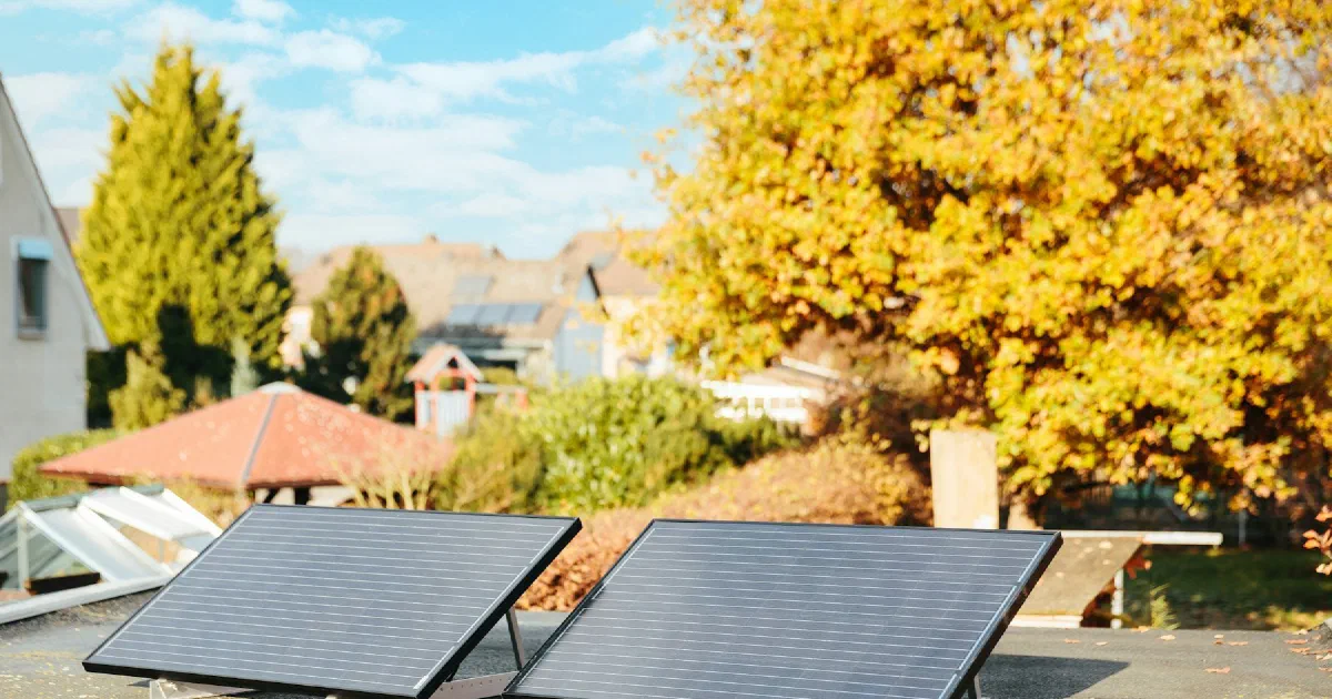 Two solar panels on a flat rooftop with a house and bright autumn trees behind.