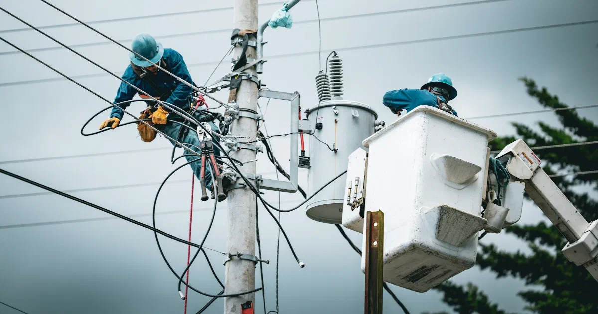 Two line workers in hard hats repairing equipment on a utility pole from a bucket truck.