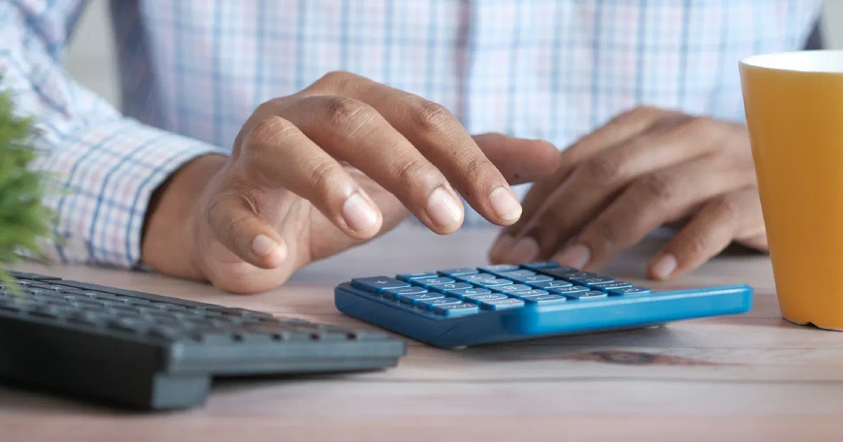 Hands typing on a blue calculator at a desk beside a keyboard and a yellow mug.