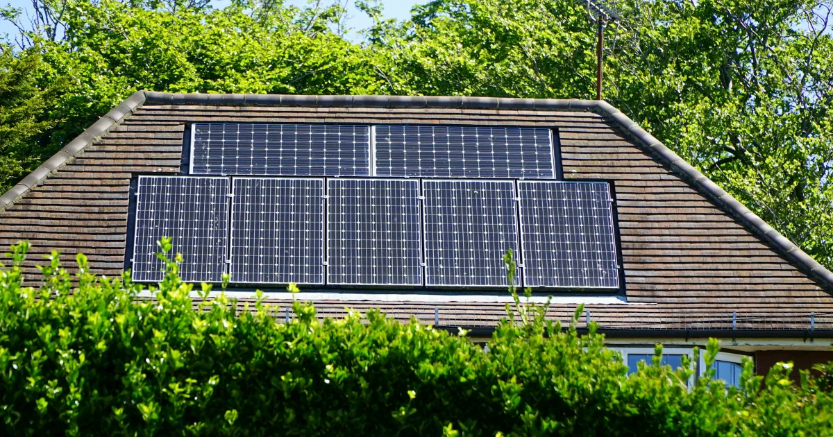 Dark solar panels mounted on a brown tiled roof behind a green hedge.