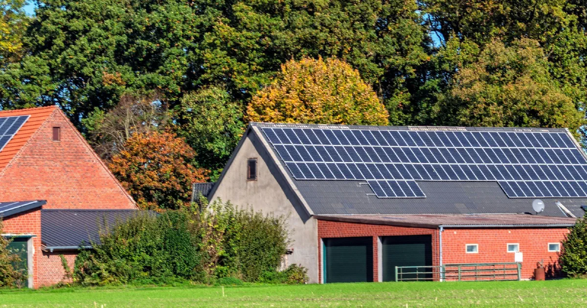 Brick barn with a roof covered in solar panels beside a green lawn and trees.